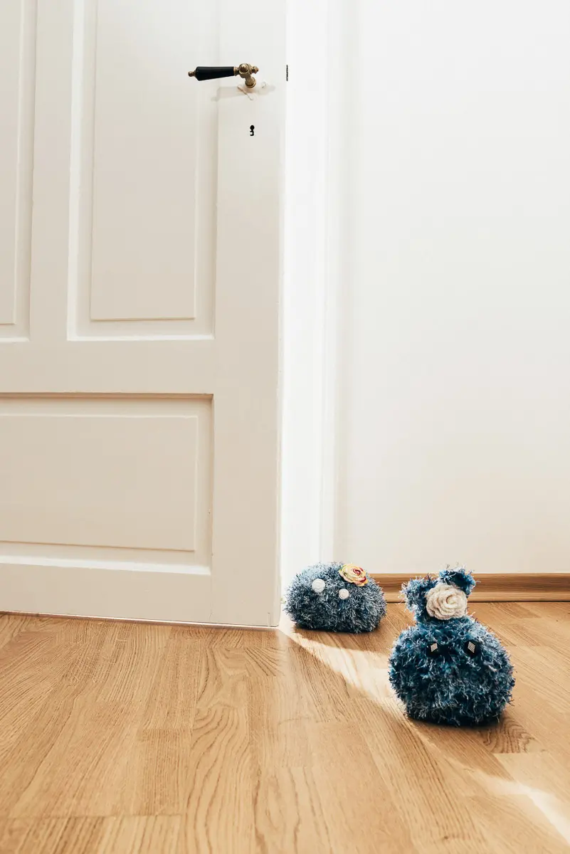 Zwei blaue Flauschwesen mit hellen Knopfaugen sitzen vor einer weißen Kassettentür auf Eichenparkett