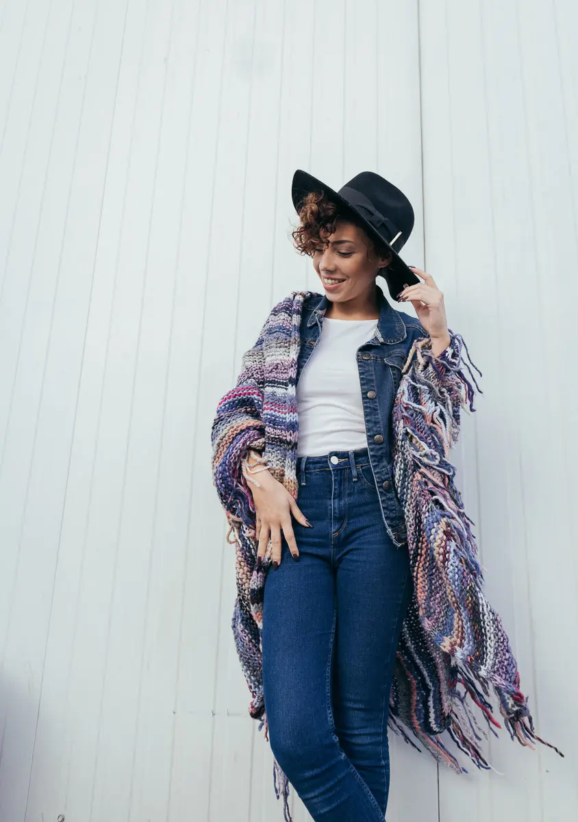 Model mit schwarzem Hut trägt bunte Fransenstola über Jeansjacke vor weißer Holzwand