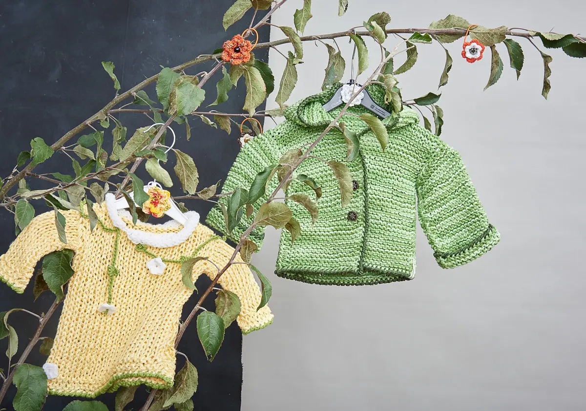 Gelbes Babyshirt und grüne Kapuzenjacke aus krausem Strick auf Kleiderbügeln zwischen Blättern