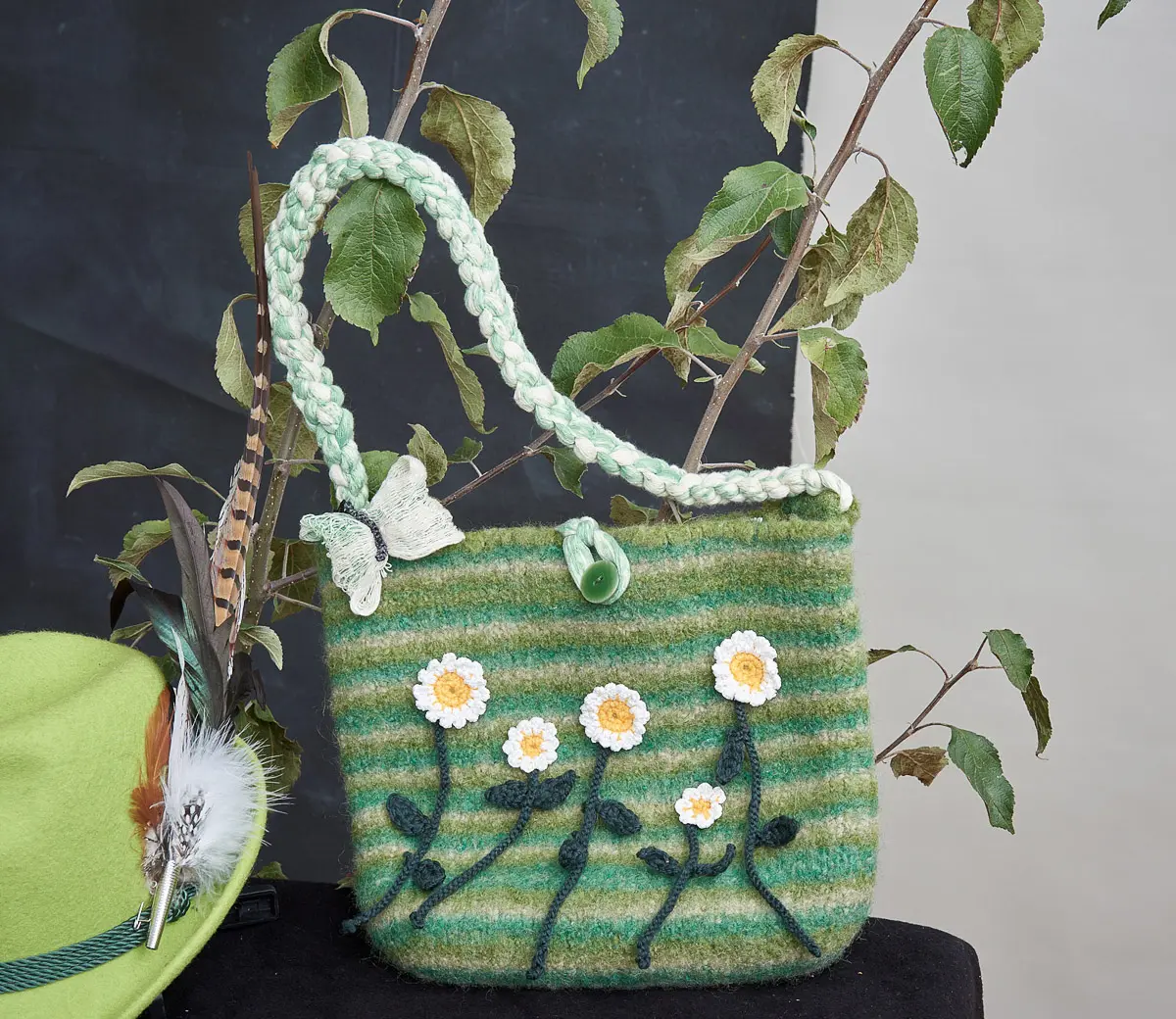Grün gestreifte Filztasche mit gehäkelten Margeriten und geflochtenem Henkel neben einem hellgrünen Hut