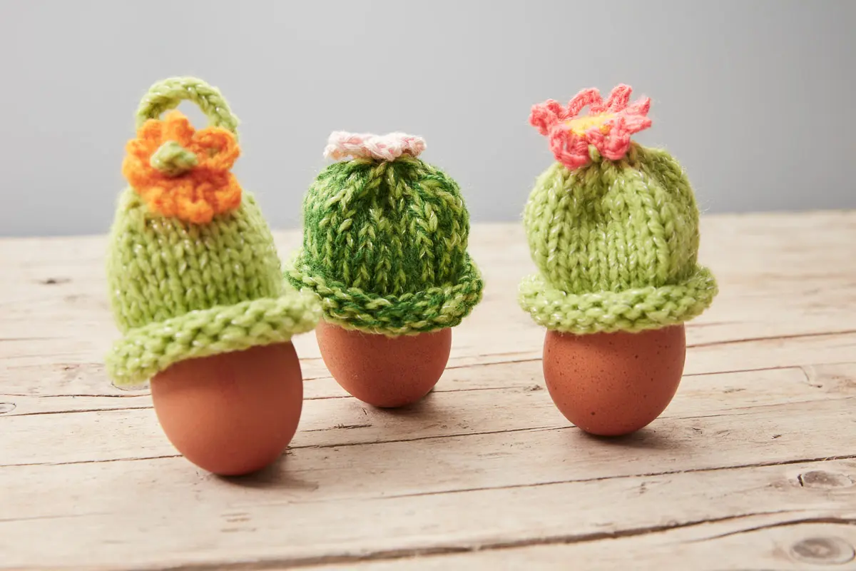 Drei grüne Eierwärmer mit gehäkelten Blüten in Orange, Rosa und Koralle auf der Spitze