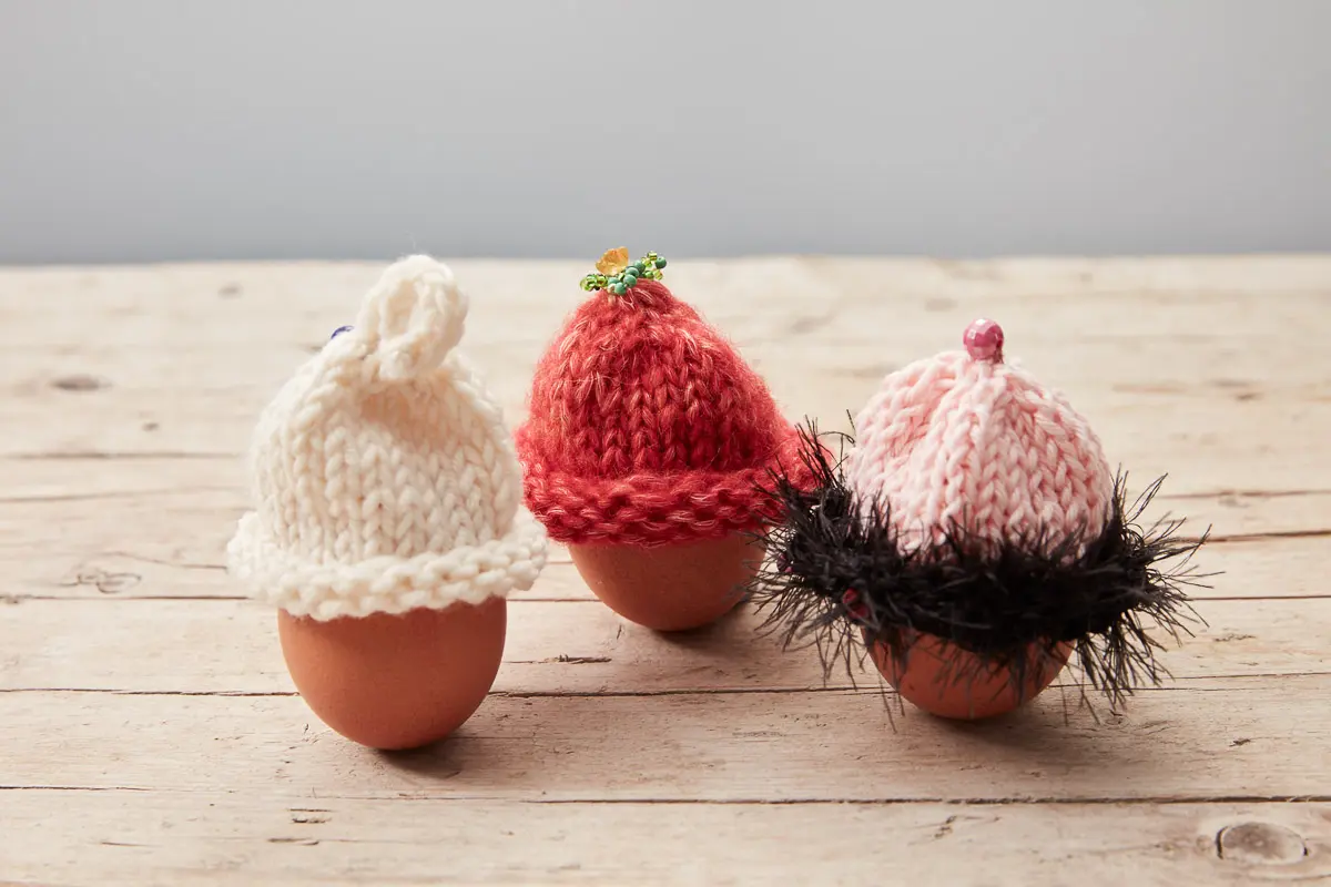 Cremefarbener, roter und rosa Eierwärmer, letzterer mit flauschigem schwarzem Fransenrand und kleiner Perlenspitze