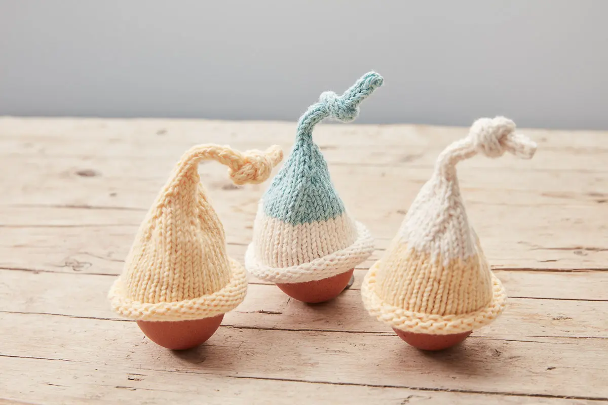 Drei Eierwärmer in Creme, Vanillegelb und Mint mit geknoteter Zipfelspitze auf einem Holztisch