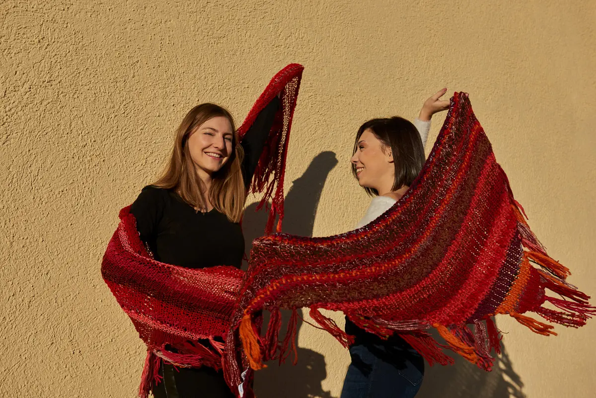Zwei Personen halten lachend rot-orange Fransentücher wie Flügel in die tiefstehende Sonne