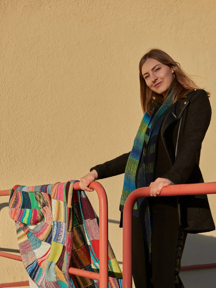 Model mit blau-grün gestreifter Stola am Geländer, daneben eine Patchwork-Decke aus Strickquadraten