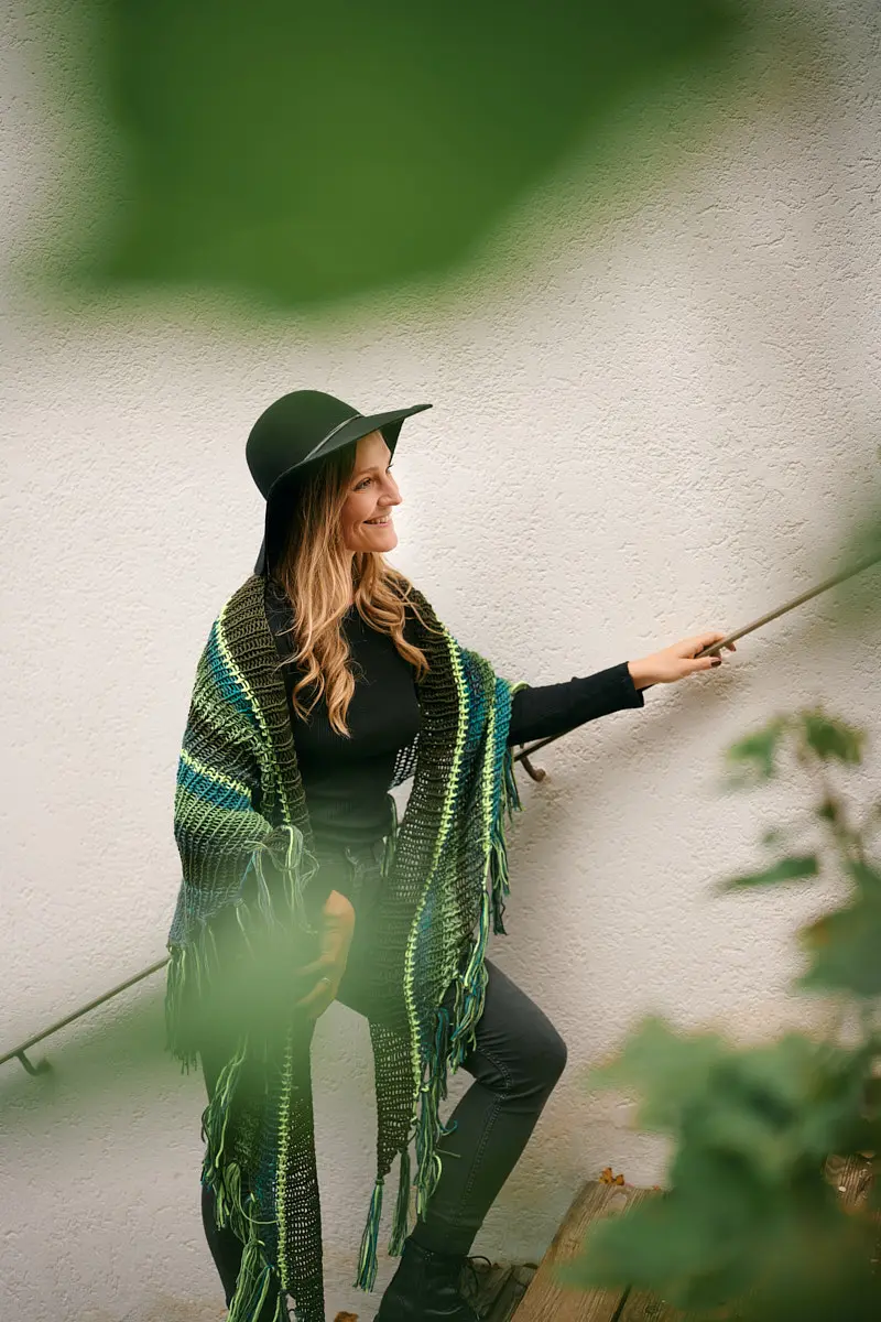 Model mit schwarzem Schlapphut und grün-türkisem Fransenponcho an einem Treppengeländer im Grünen