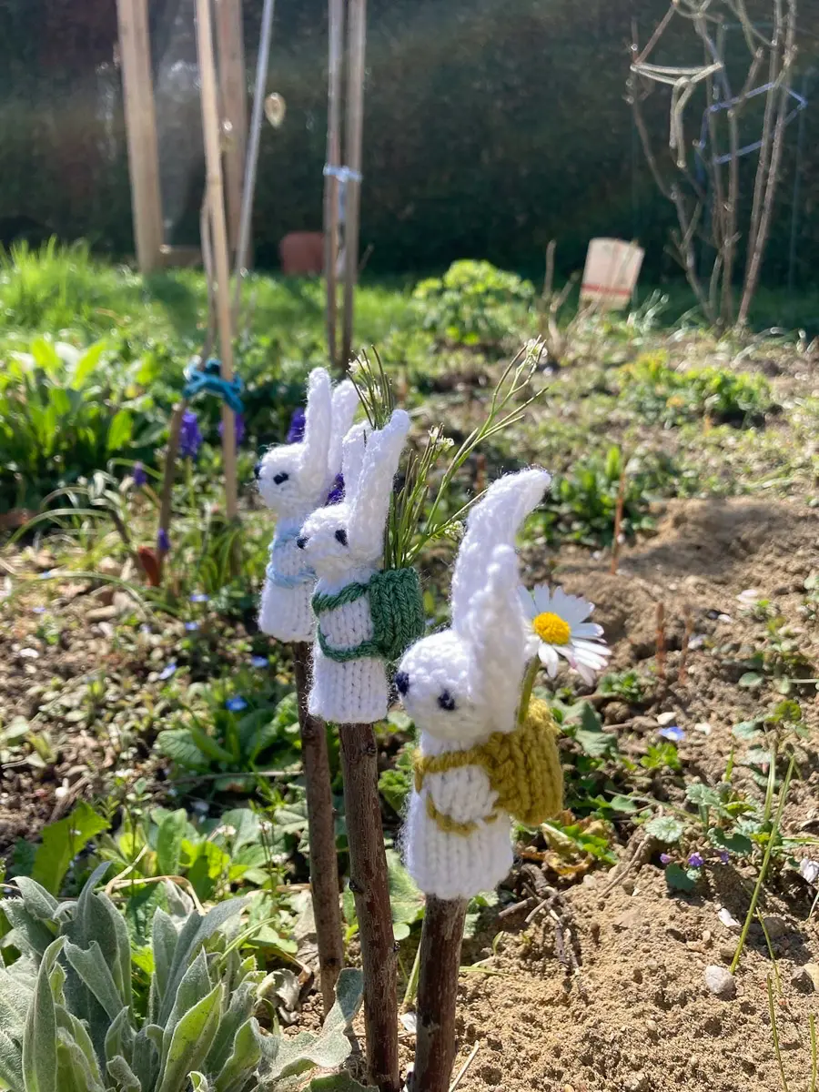 Drei kleine Strickhasen auf Aststäben in einem sonnigen Gartenbeet, einer mit senfgelbem Täschchen