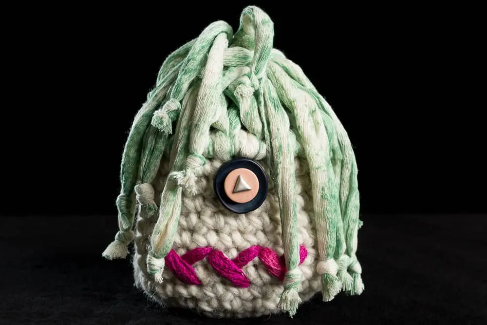 Wollweiße Figur mit mintgrünen Fransensträhnen, einem einzelnen Knopfauge und magentafarbenem Stickmund